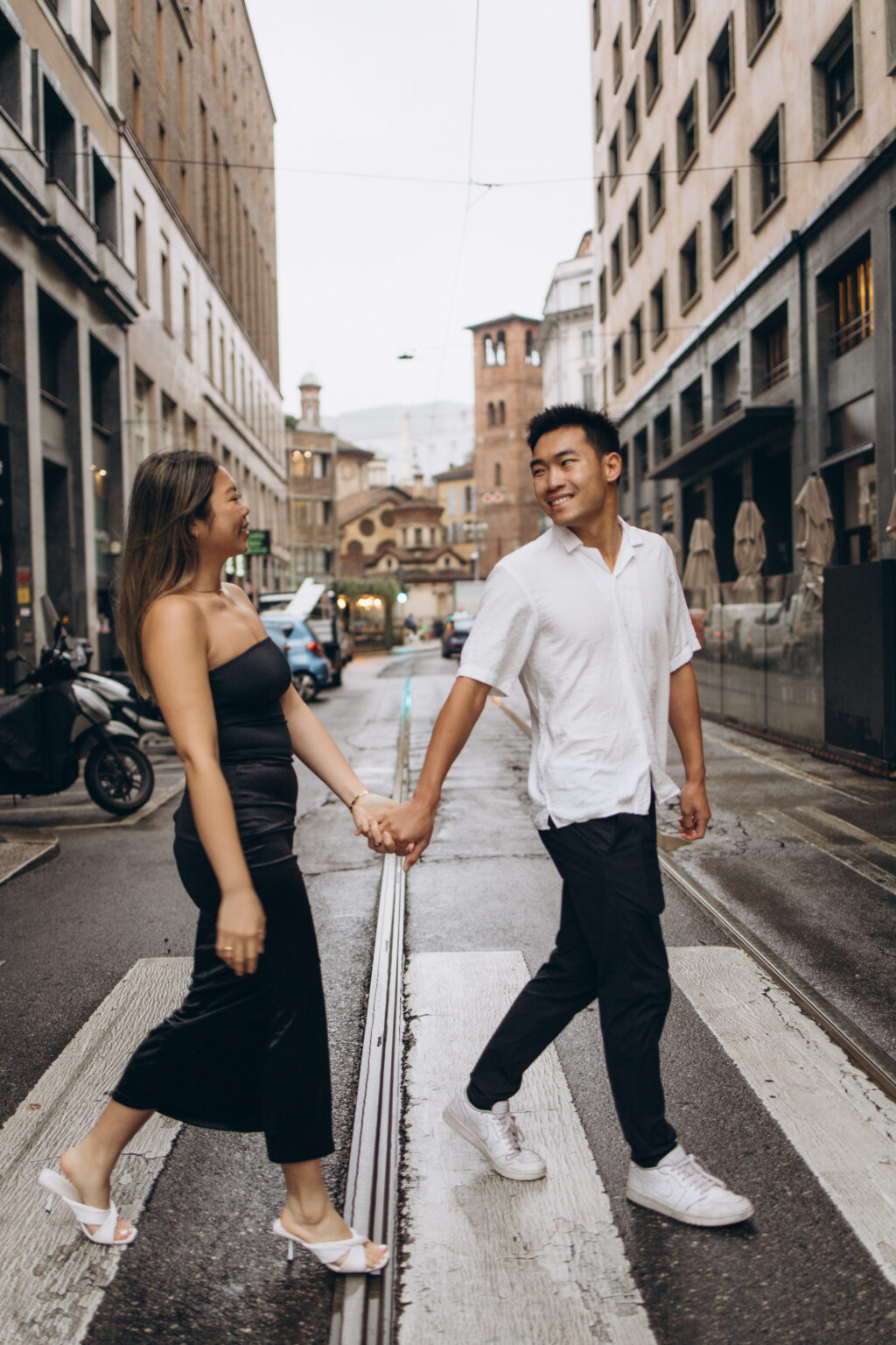 A couple holding hands and crossing a Milan street on a crosswalk, smiling at each other with a bell tower behind them