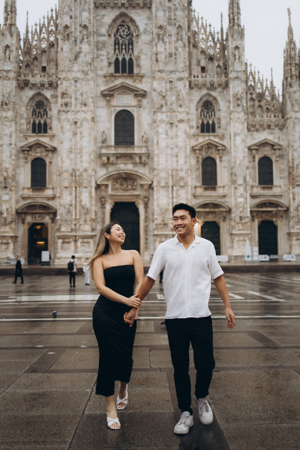 A smiling couple walking hand in hand across Piazza del Duomo, with the ornate facade of Milan Cathedral behind them