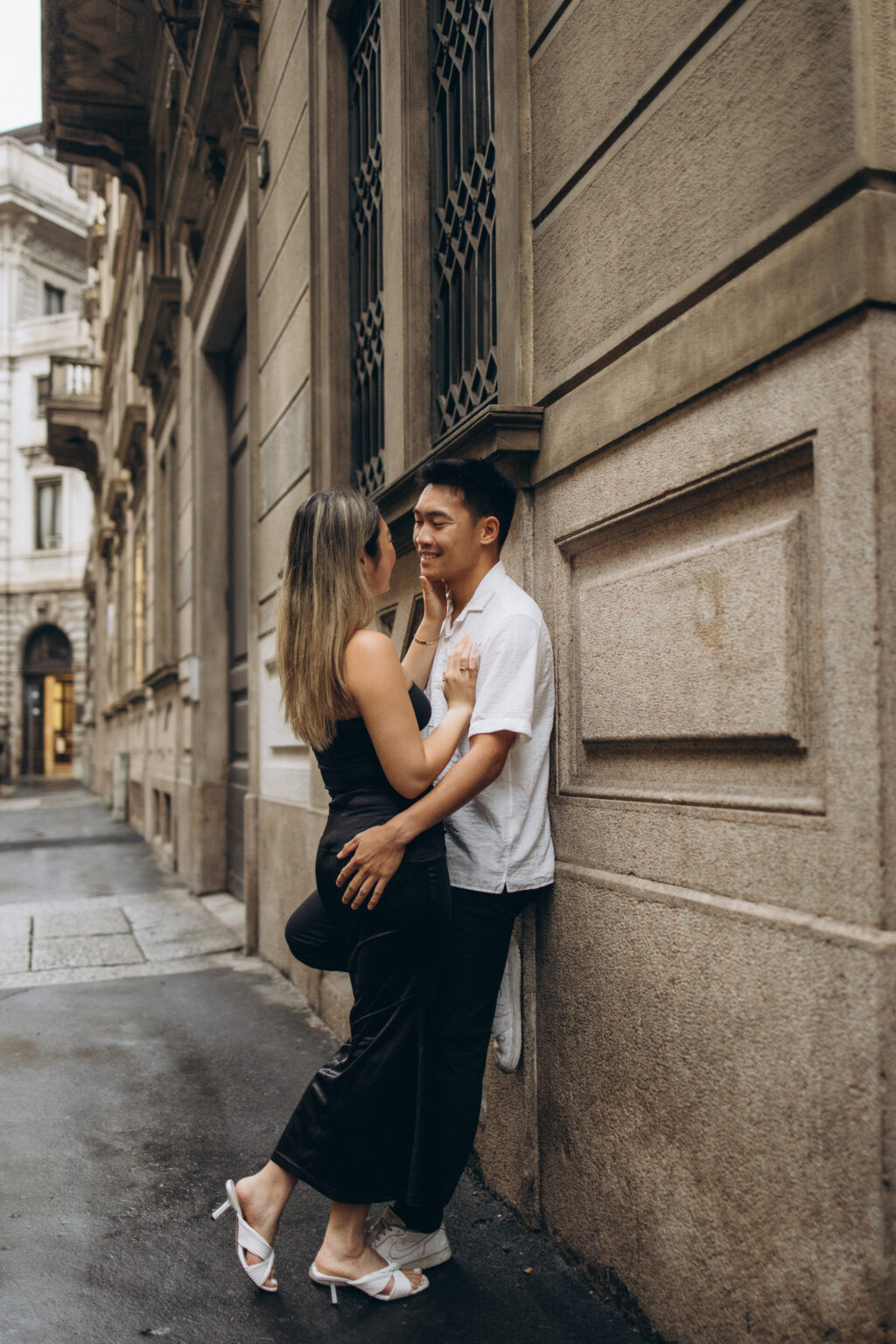 A couple in an intimate embrace on a quiet Milan street, leaning against an elegant stone building facade