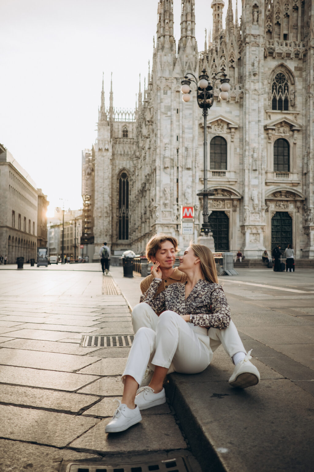 A couple sitting close together in Piazza del Duomo at sunrise, with Milan Cathedral rising behind them