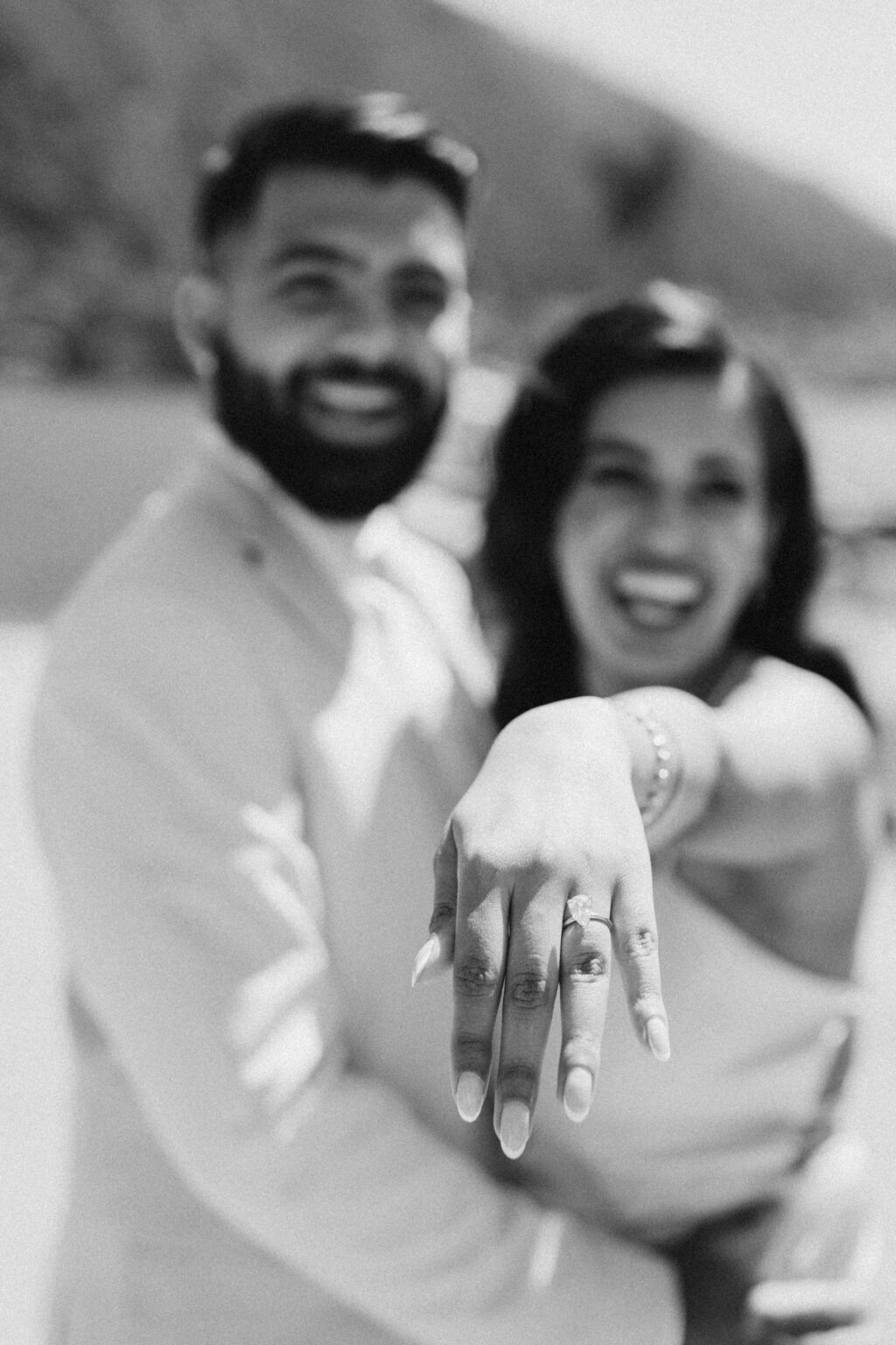 A black and white portrait of a laughing newly engaged couple with the bride-to-be reaching her ring hand toward the camera, on Lake Como.