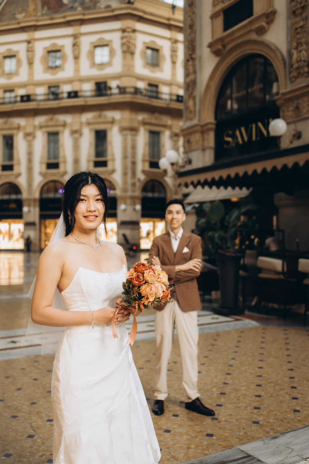 Smiling bride-to-be holding a peach rose bouquet with her groom-to-be standing proudly behind her in the Galleria Vittorio Emanuele II in Milan