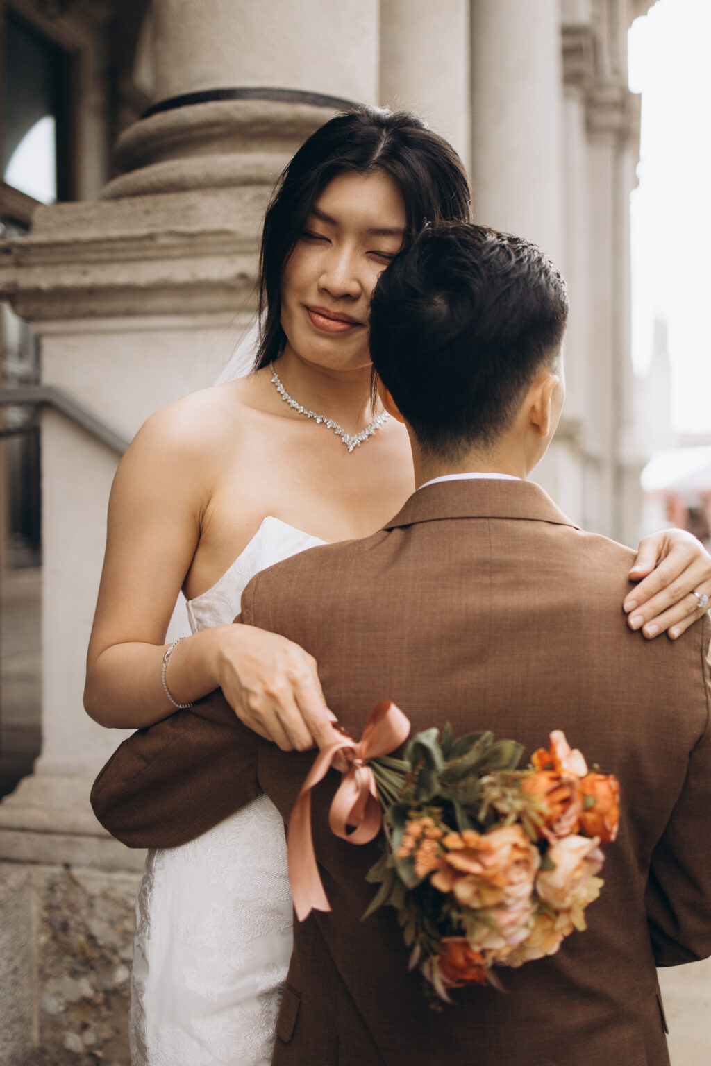 Bride-to-be with eyes closed embracing her groom-to-be with her arms around his neck and bouquet in hand among the columns of Piazza dei Mercanti in Milan