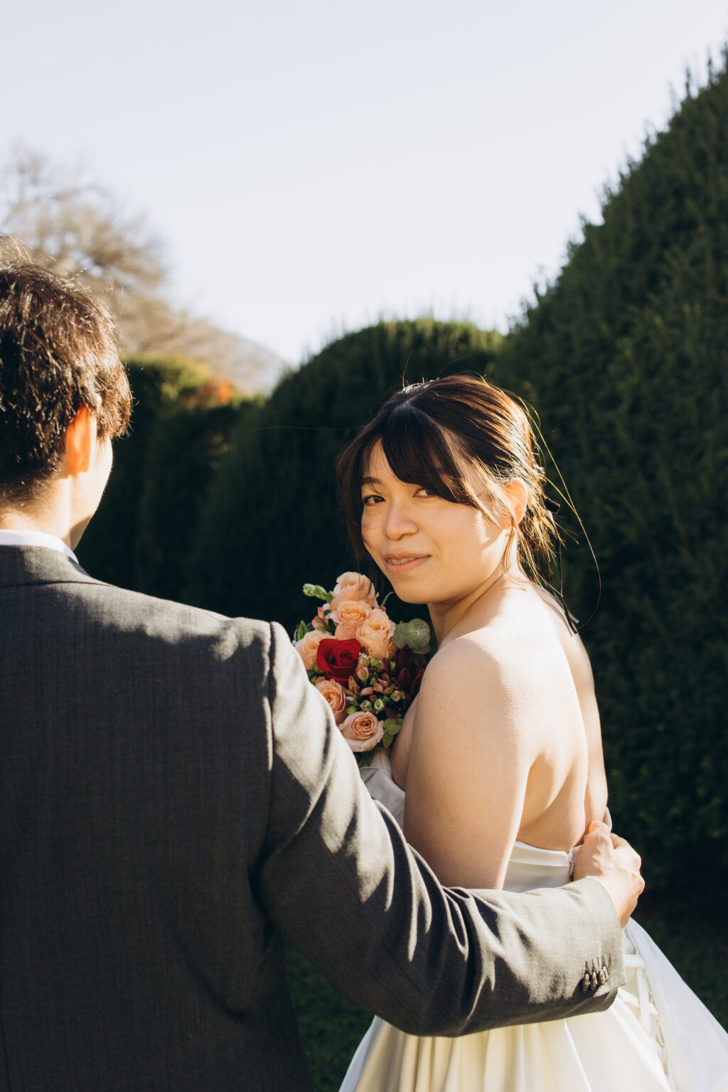 Bride glancing back over her bare shoulder with a bouquet while her groom holds her in a topiary garden on Lake Como