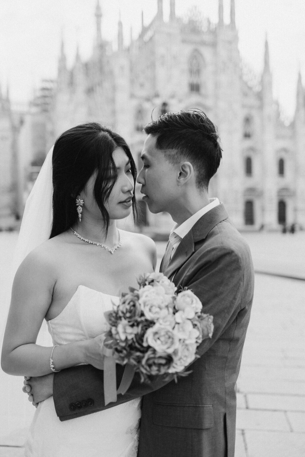 Bride-to-be and groom-to-be leaning close together with a bouquet in front of the Duomo in Milan, black and white