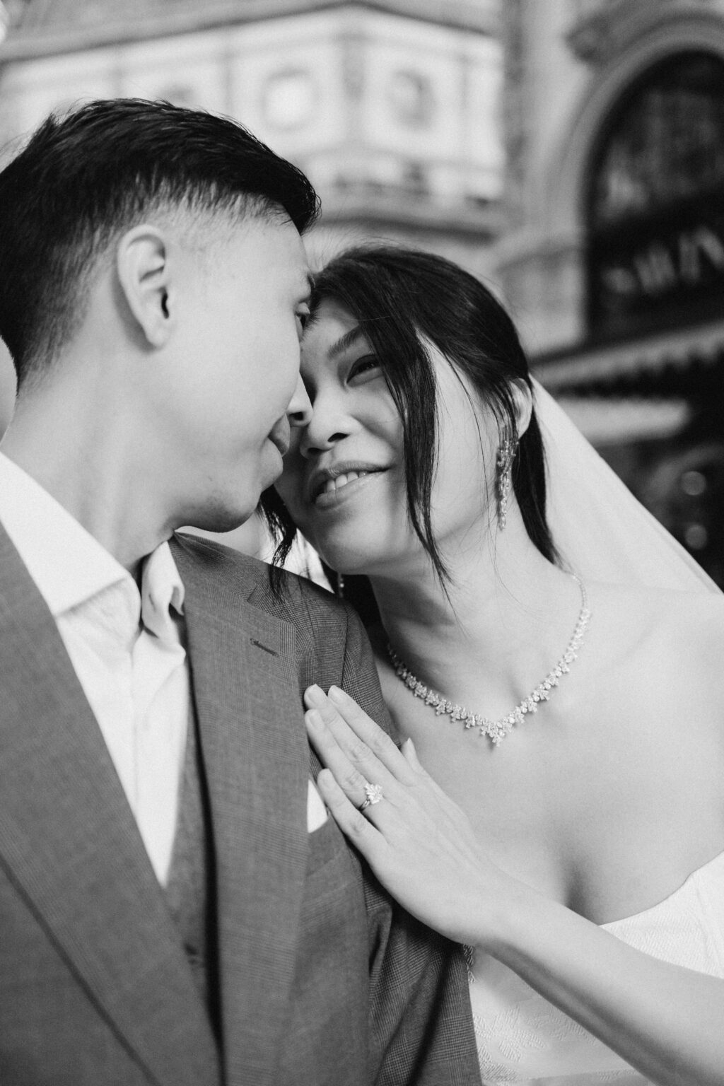 Bride-to-be resting her hand on her groom-to-be's chest as they lean close together, her ring catching the light, in the Galleria in Milan, black and white