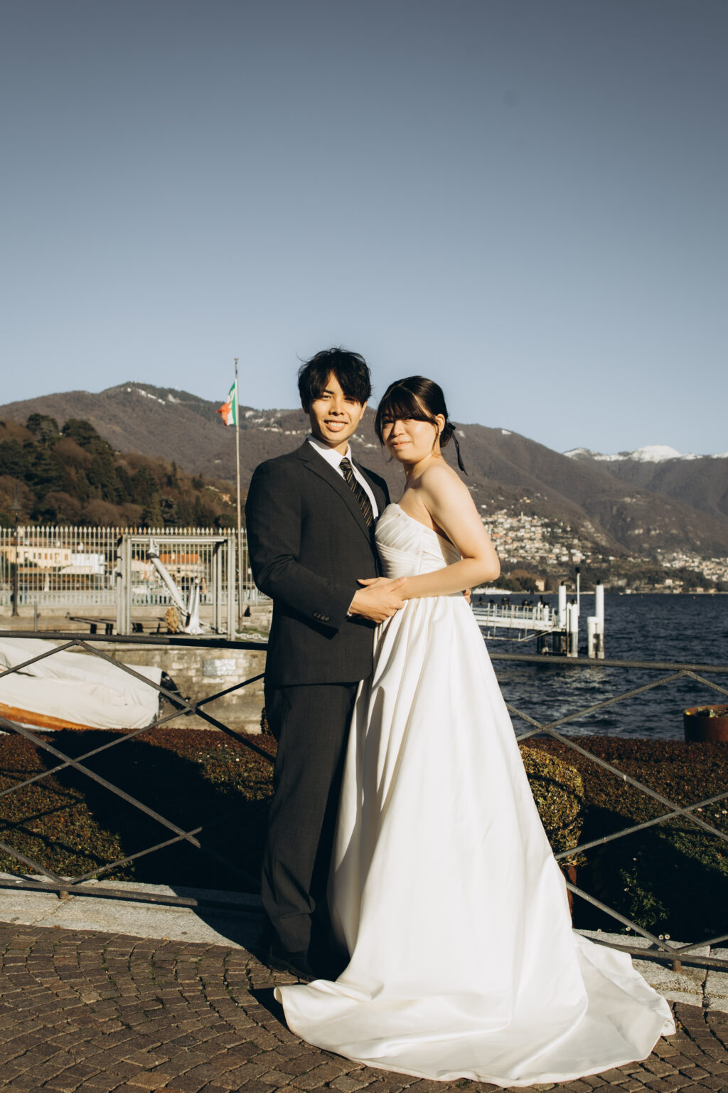 Bride and groom posing together at a waterfront railing on Lake Como with the Italian flag, jetty and mountains behind them