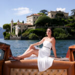 Woman in a white dress reclining barefoot on the bow of a wooden boat with Villa del Balbianello behind her on Lake Como