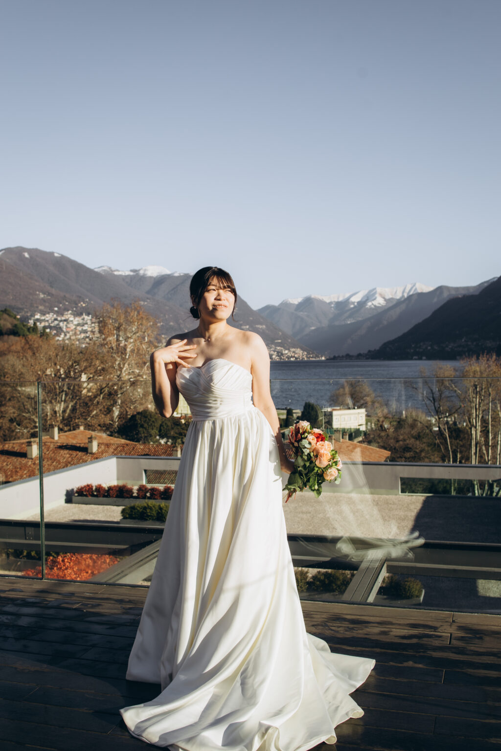 Bride in a strapless gown holding a bouquet on a rooftop terrace with her veil drifting and snow-capped mountains behind her over Lake Como