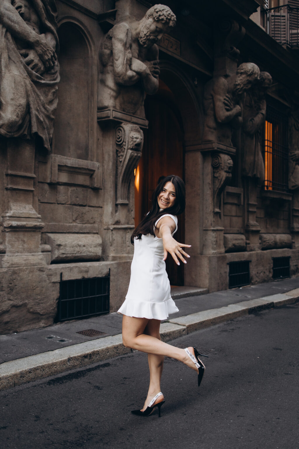 Woman in a short white dress posing playfully in front of the telamon statues of the Casa degli Omenoni on Via degli Omenoni in Milan