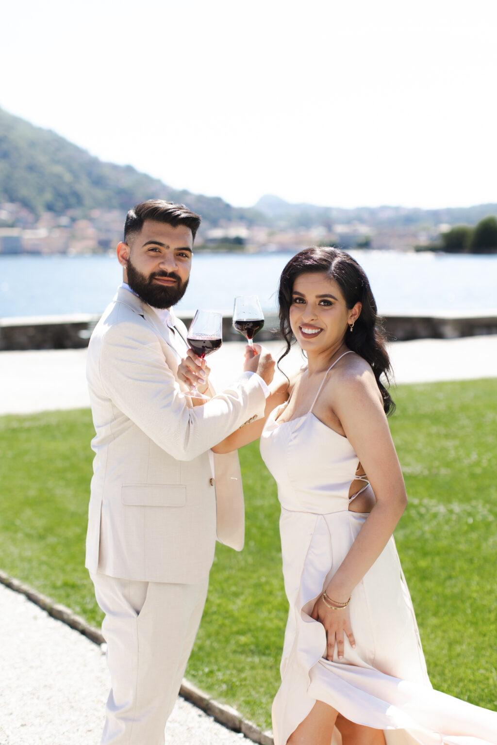 A newly engaged couple raising red wine glasses to celebrate on a green lakeside lawn on Lake Como.