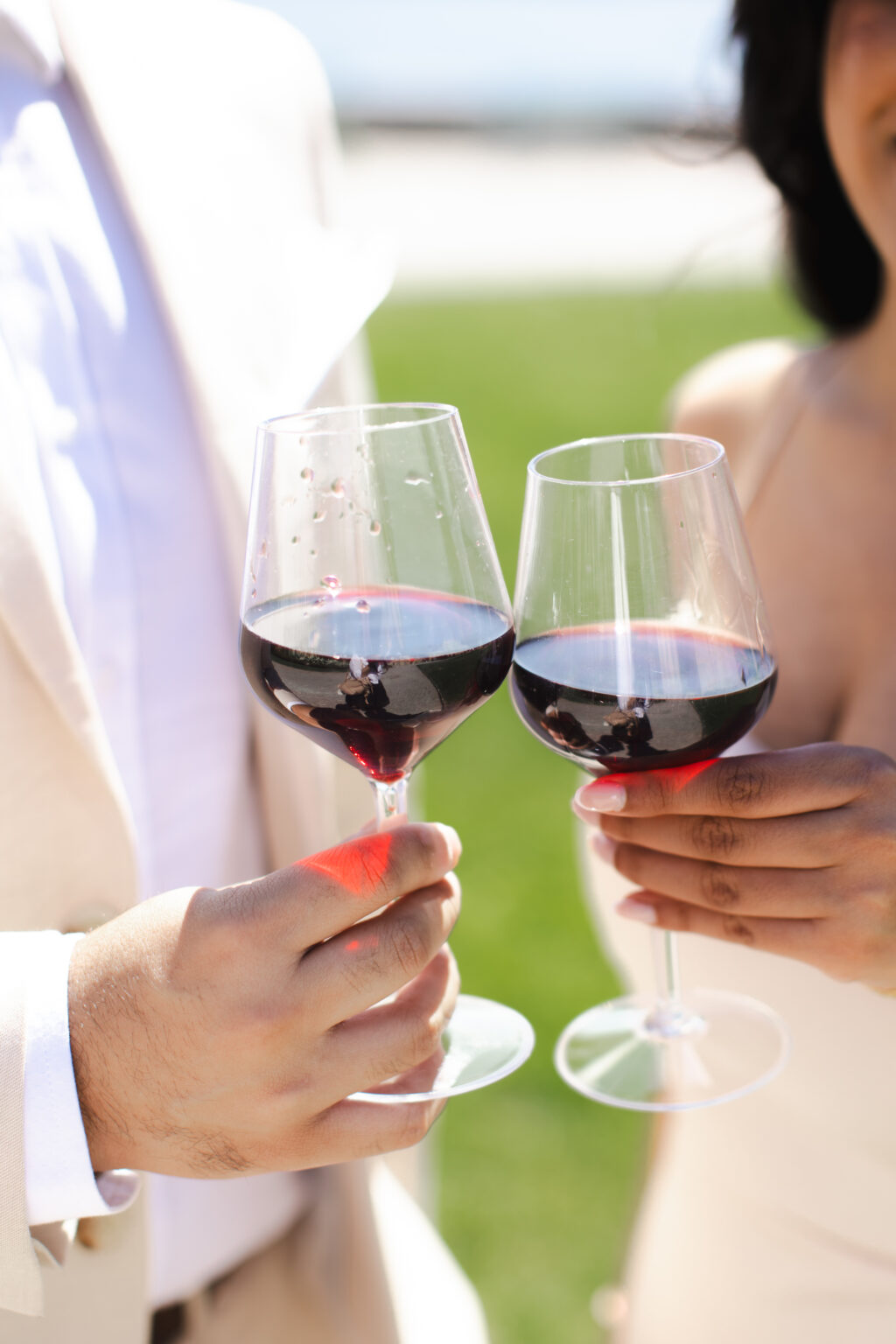 A close-up of two red wine glasses clinking in an engagement toast, held by a newly engaged couple on a green lakeside lawn on Lake Como.