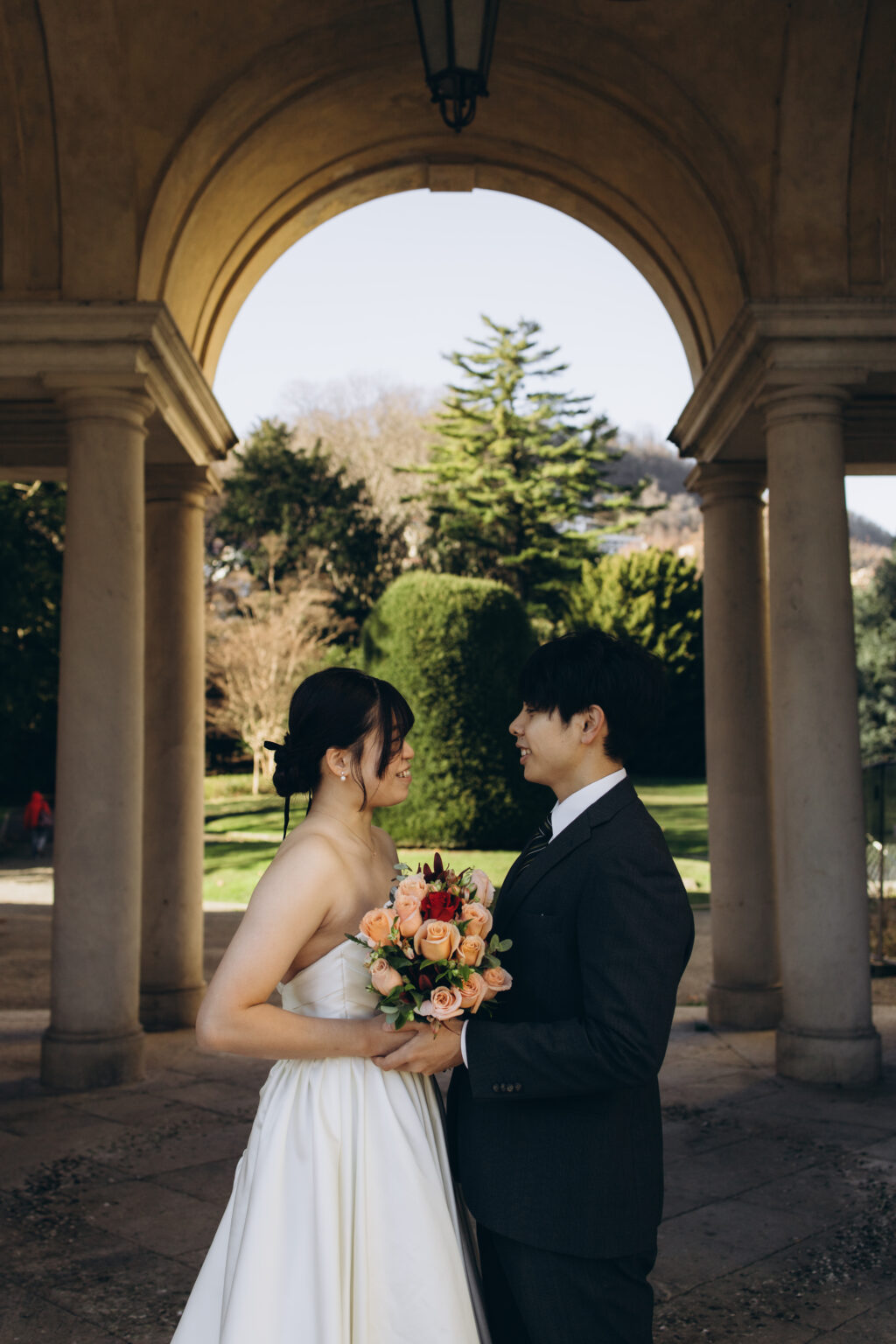 Bride and groom smiling at each other while holding her bouquet beneath a stone arch opening onto a garden on Lake Como