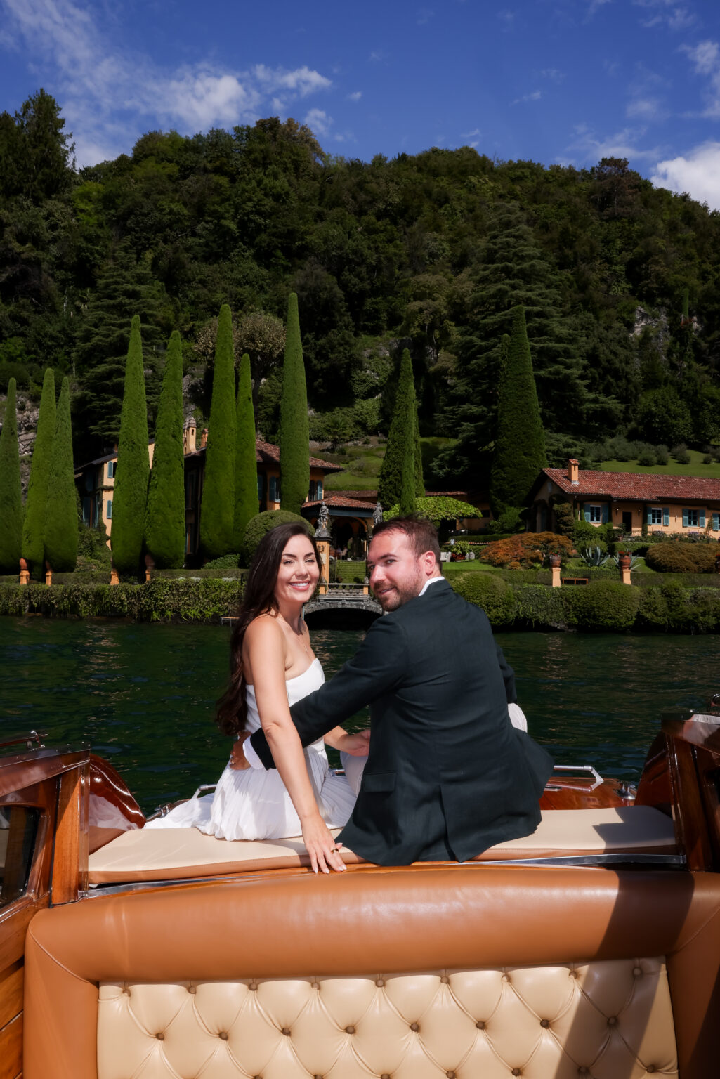Couple turning to smile over their shoulders while seated on a wooden boat, with a cypress-lined lakeside villa garden behind them on Lake Como