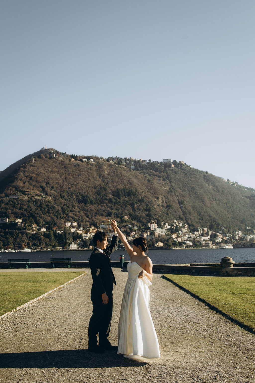 Groom twirling his bride on the lakefront lawn of Como with Monte Brunate and the town rising across the water behind them