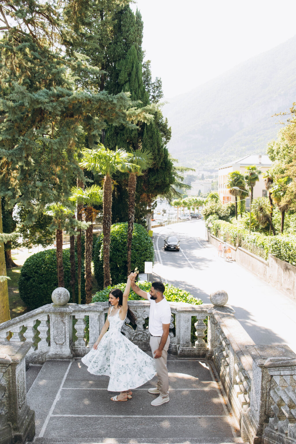 Newly engaged couple twirling at the top of the Parco Teresio Olivelli staircase above the lakeside road in Tremezzo, surrounded by palms and cypresses