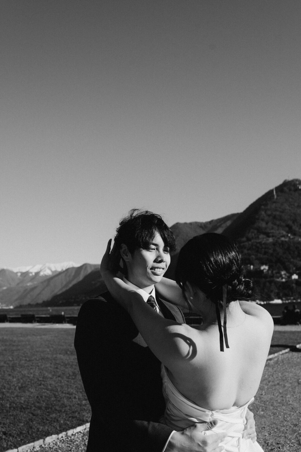 Bride cradling her groom's face in an embrace on the Como lakefront with snow-capped mountains behind them, black and white