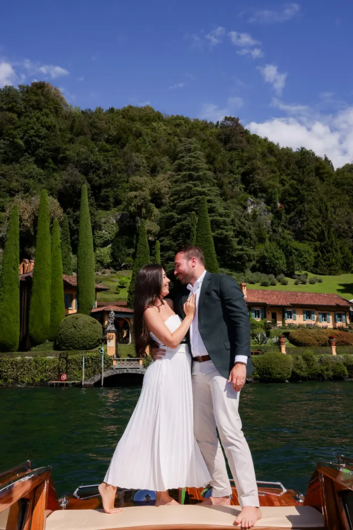 Couple embracing on the bow of a wooden boat in front of a villa with cypress trees, Lake Como
