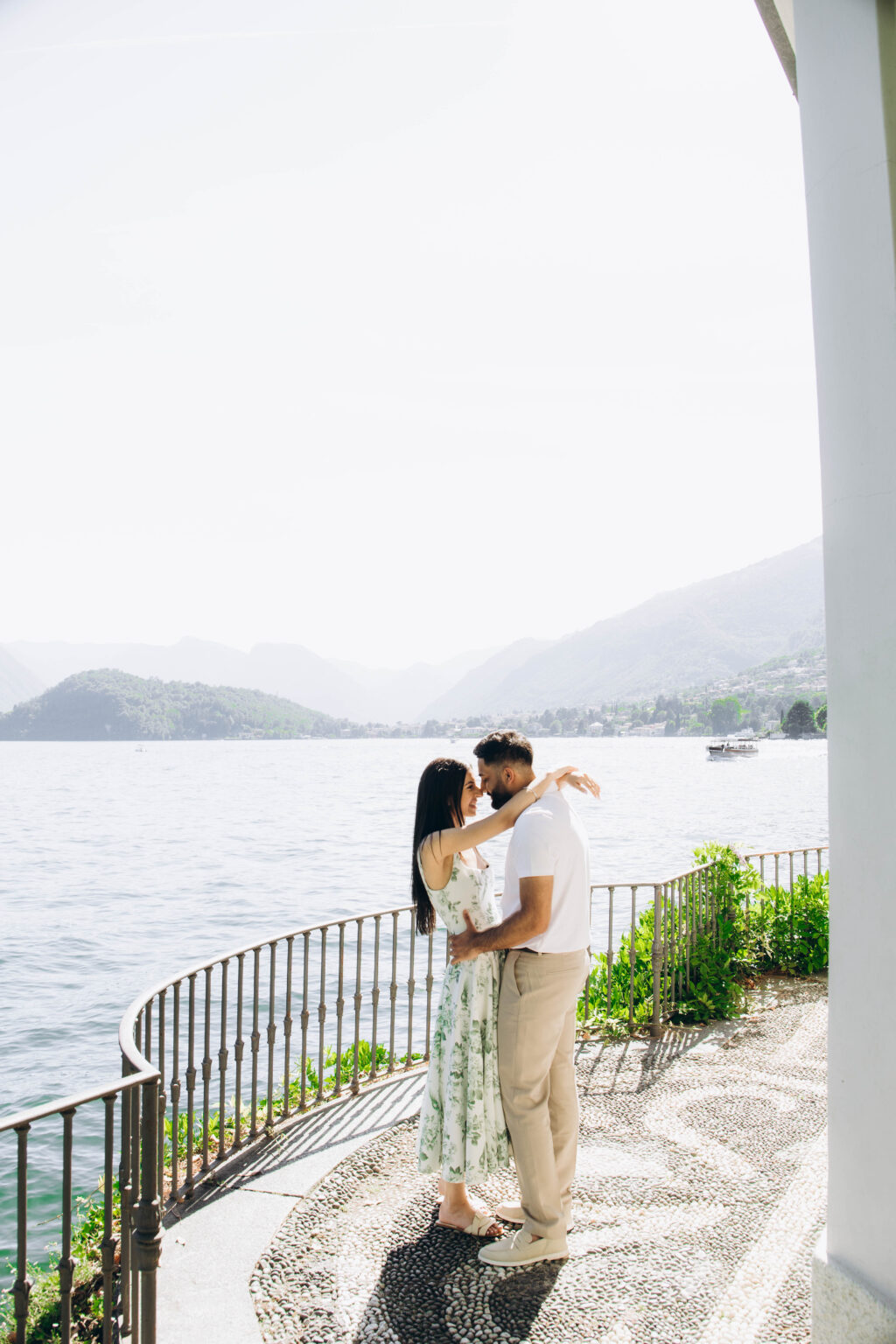 Couple sharing a forehead-to-forehead embrace on a pebble mosaic walkway beside an iron railing, with the water and mountains of Lake Como behind