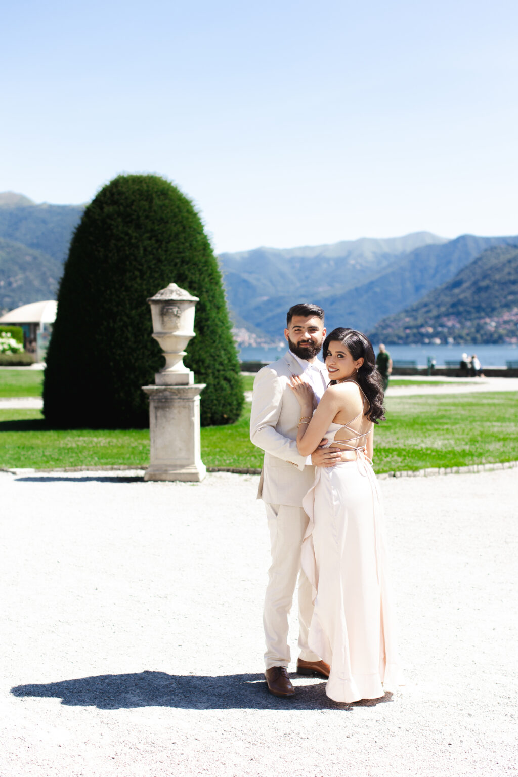 Couple embracing beside a tall topiary and a stone garden urn in a villa garden on Lake Como with mountains behind them