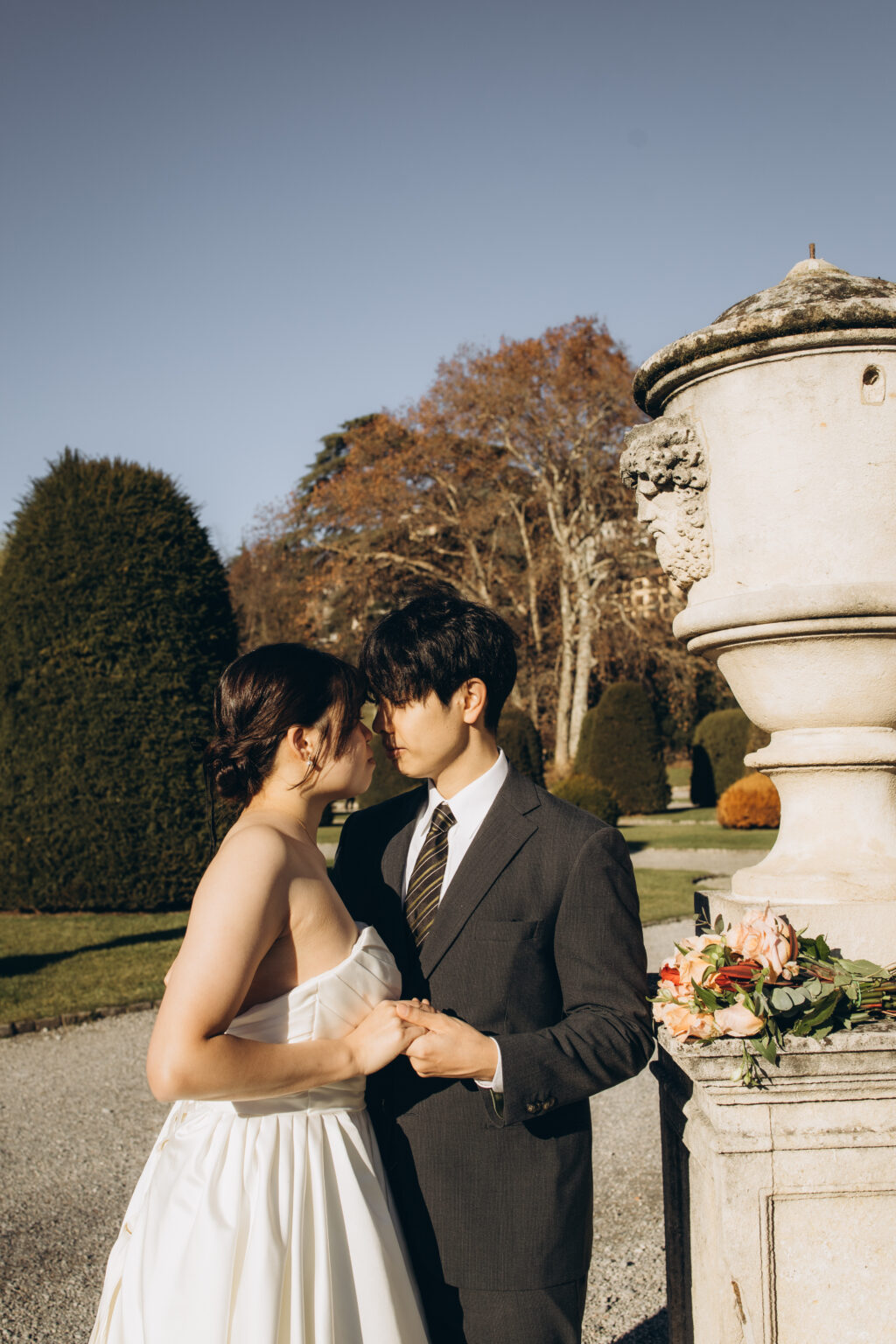 Bride and groom leaning forehead to forehead and holding hands beside a stone urn in a villa garden on Lake Como