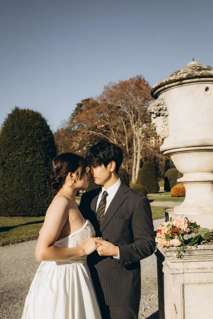 Bride and groom leaning forehead to forehead and holding hands beside a stone urn in a villa garden on Lake Como