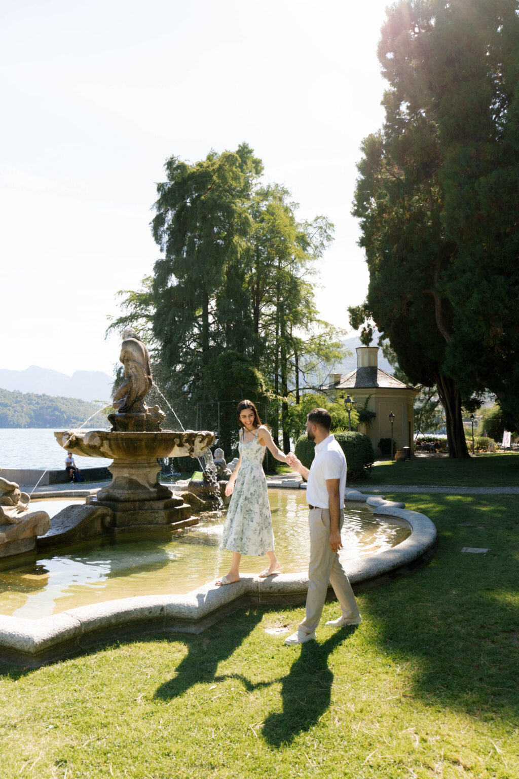 Woman walking along the rim of the central fountain in Parco Teresio Olivelli as her partner holds her hand, with the octagonal Tarocchiera pavilion and Lake Como behind