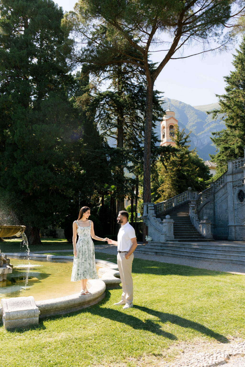 Couple holding hands at the fountain in Parco Teresio Olivelli, with the monumental staircase, umbrella pines, a church bell tower, and mountains in the background