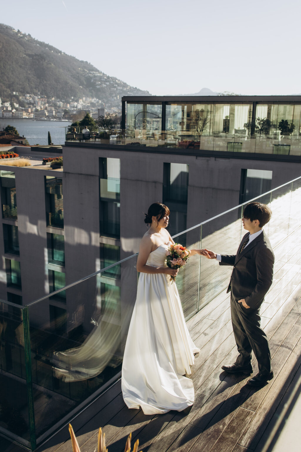 Bride and groom holding hands along a glass-railed terrace of a modern lakeside hotel overlooking Lake Como and the town of Como