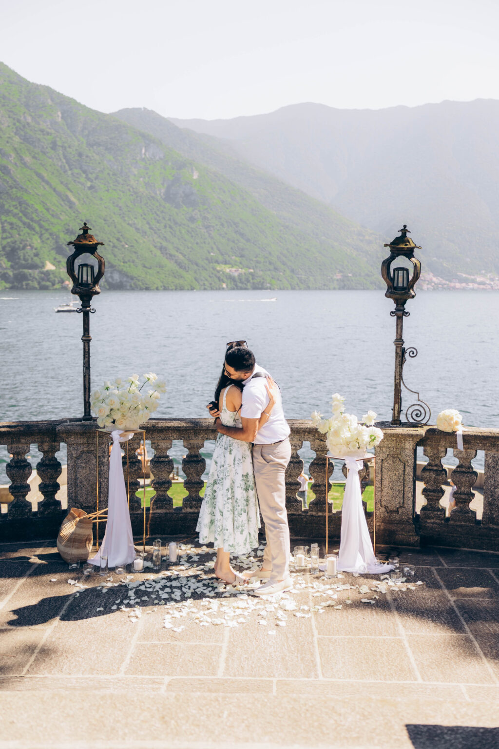 Newly engaged couple in a tight embrace on the petal-strewn proposal terrace between white rose arrangements and antique lanterns, Lake Como behind