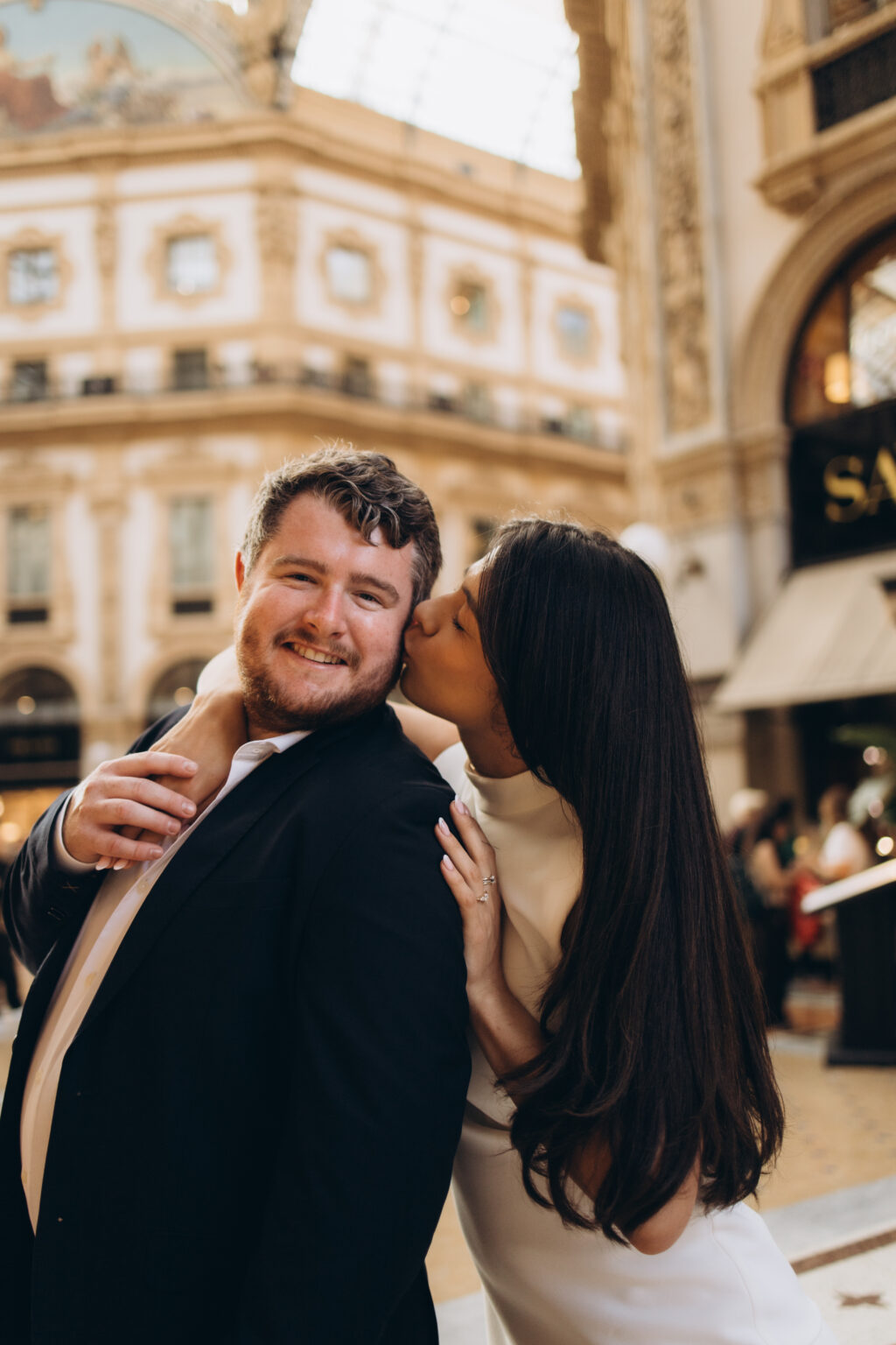Woman kissing her smiling fiance on the cheek beneath the glass dome of the Galleria Vittorio Emanuele II in Milan