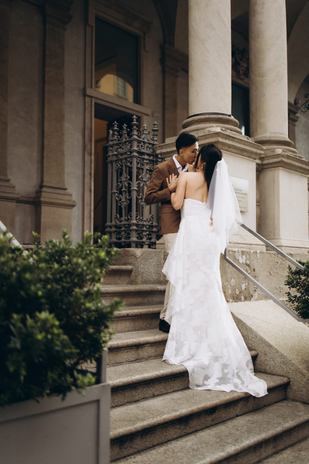 Bride-to-be and groom-to-be kissing on the stone steps of the Palazzo Giureconsulti on Piazza dei Mercanti in Milan, seen from behind