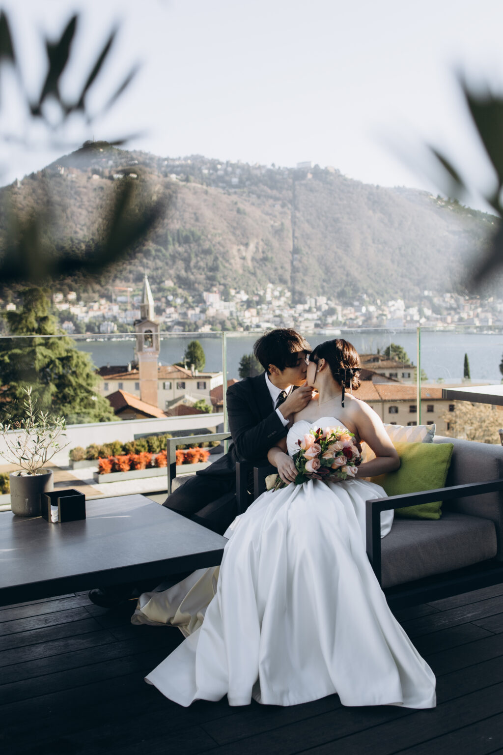 Bride and groom kissing on a terrace lounge framed by olive leaves, with Como, its bell tower and the mountains behind them