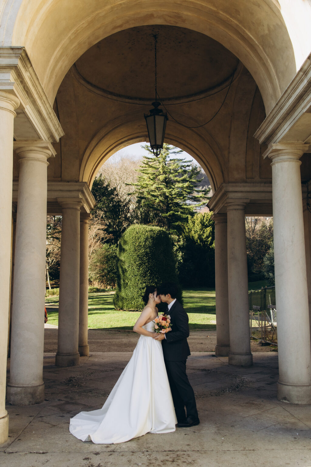 Bride and groom kissing under a grand arched colonnade opening onto a garden on Lake Como, a hanging lantern above them