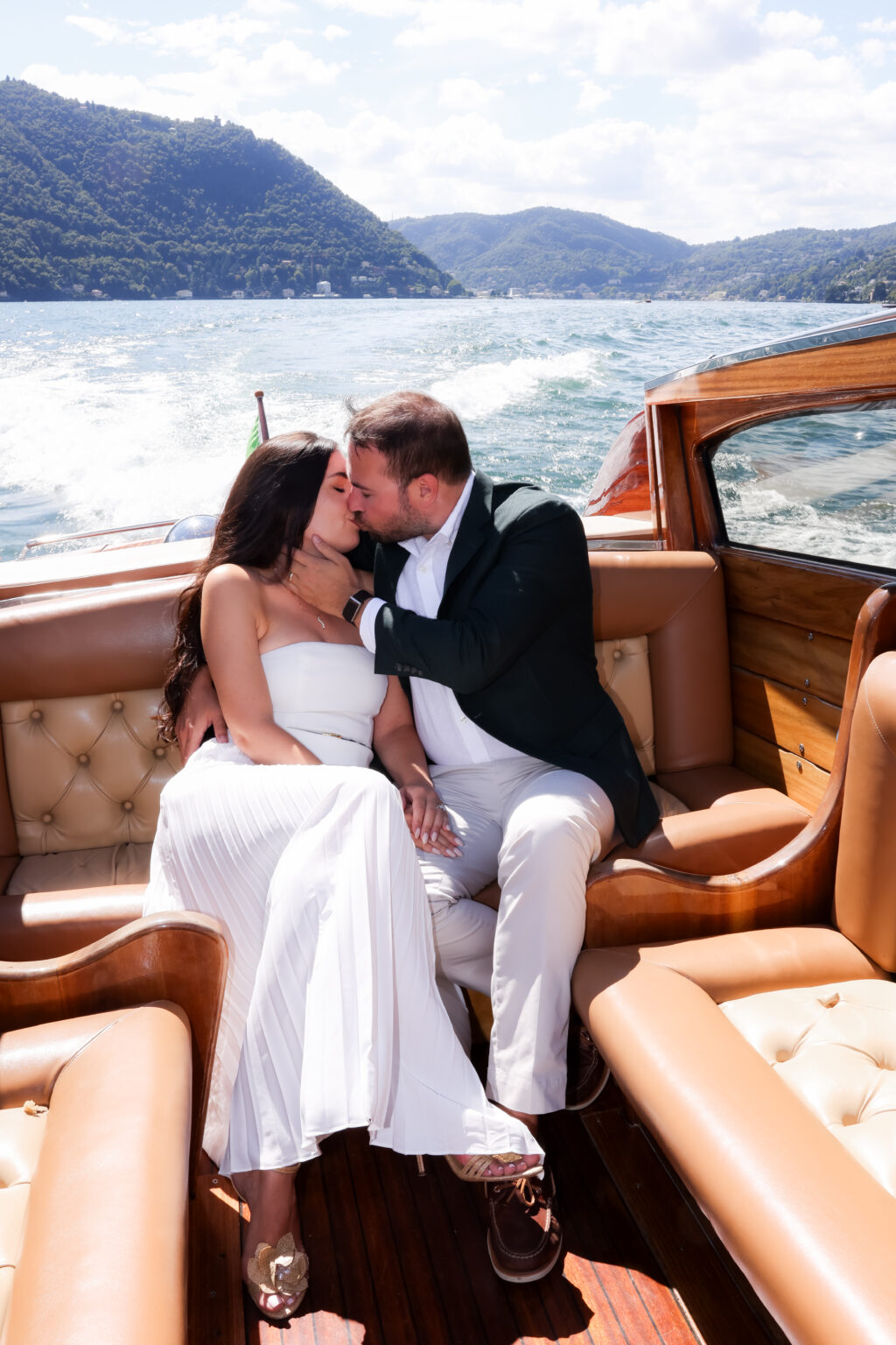 Couple sharing a kiss on the leather seats of a vintage wooden boat cruising Lake Como, mountains and open water behind them