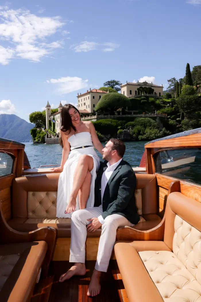 Woman laughing while sitting on the edge of a wooden boat as her partner looks up at her, Villa del Balbianello behind them on Lake Como