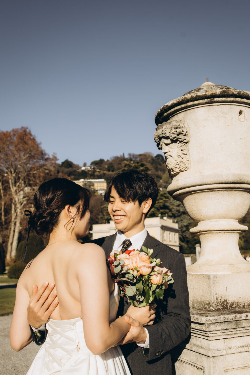 Bride and groom leaning forehead to forehead and holding hands beside a stone urn in a villa garden on Lake Como