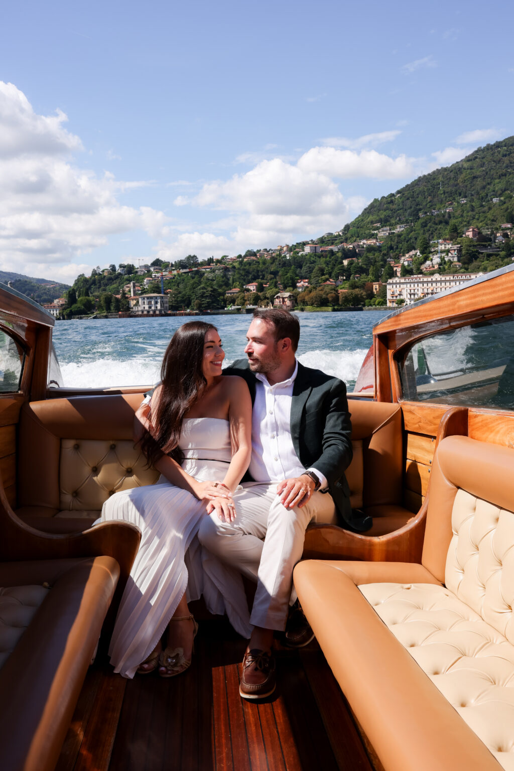 Couple sitting close together on the tan leather seats of a vintage wooden boat as it cruises Lake Como, wake and green shoreline behind them