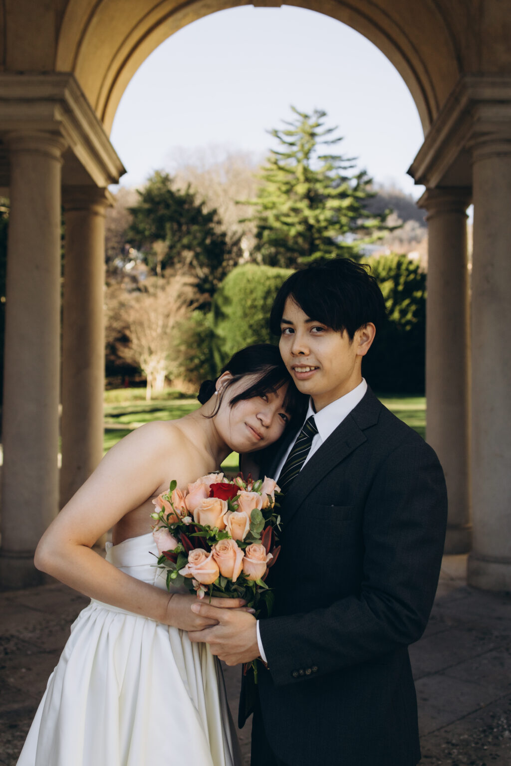 Bride resting her head on her groom's shoulder while holding her bouquet beneath a garden colonnade on Lake Como