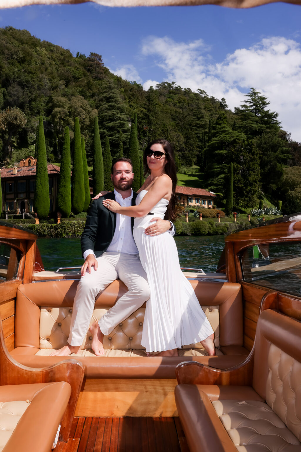 Couple posing confidently for the camera on a vintage wooden boat, the woman in sunglasses, with a cypress villa garden behind them on Lake Como