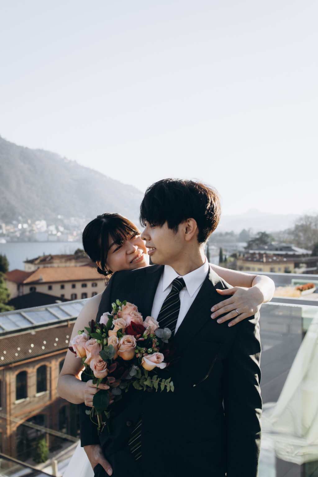 Bride embracing her groom from behind while holding a peach and burgundy bouquet on a rooftop terrace overlooking Lake Como
