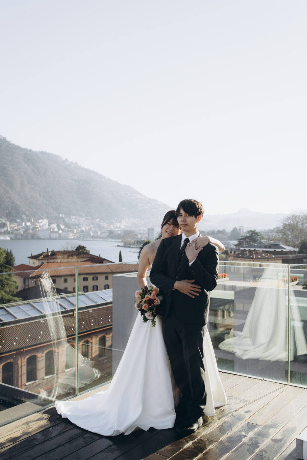 Bride embracing her groom from behind on a glass rooftop terrace overlooking Lake Como, the town and mountains beyond