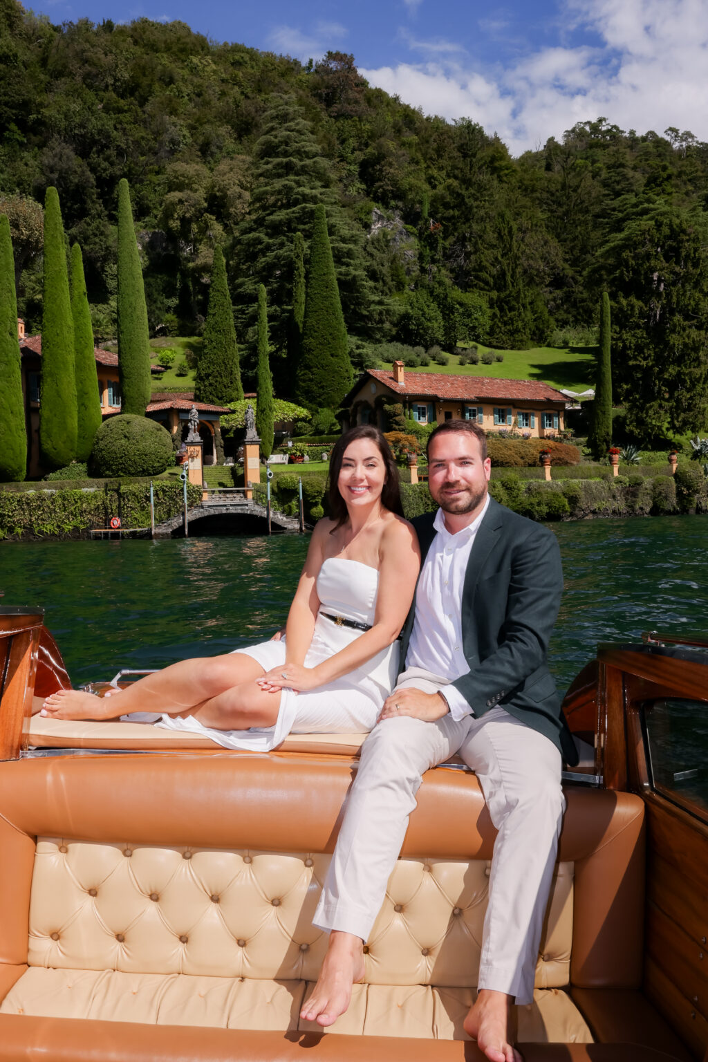 Couple sitting close together and smiling on the tan leather seat of a wooden boat, a cypress villa garden with a small stone bridge behind them on Lake Como