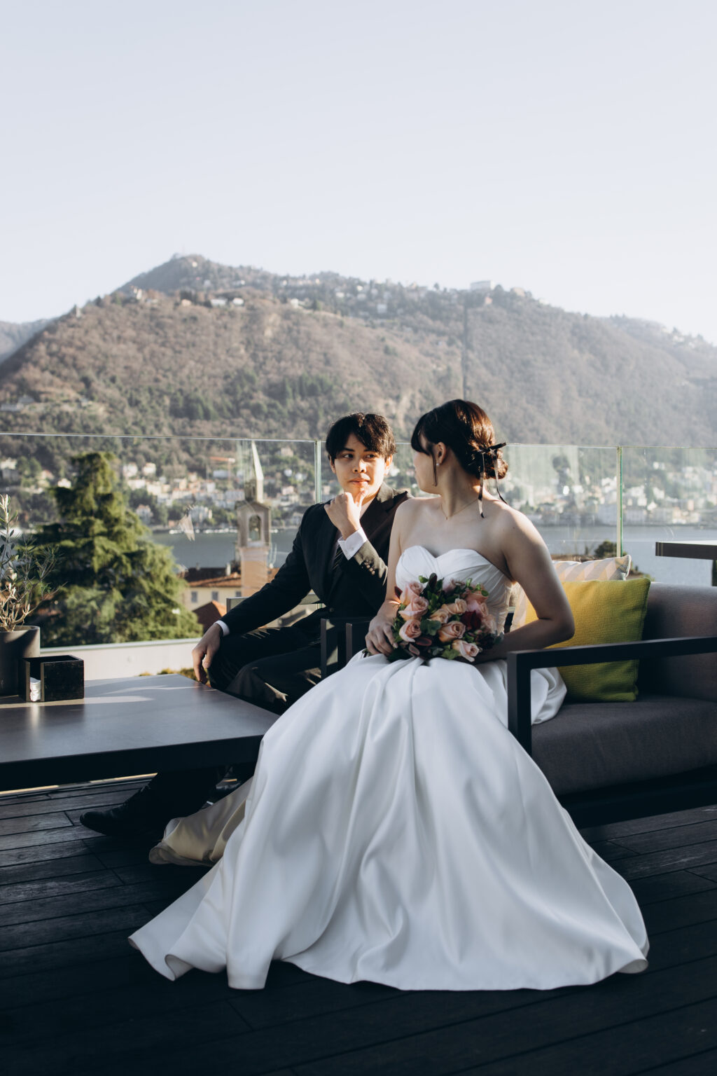 Bride and groom seated on a modern terrace lounge with the town of Como, its bell tower and Monte Brunate behind them