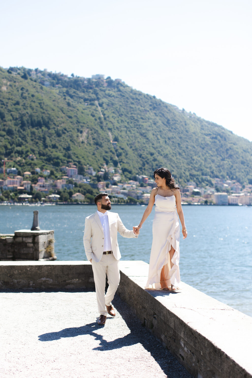 Couple holding hands and walking along a stone lakefront wall on Lake Como, the town of Como and Monte Brunate behind them