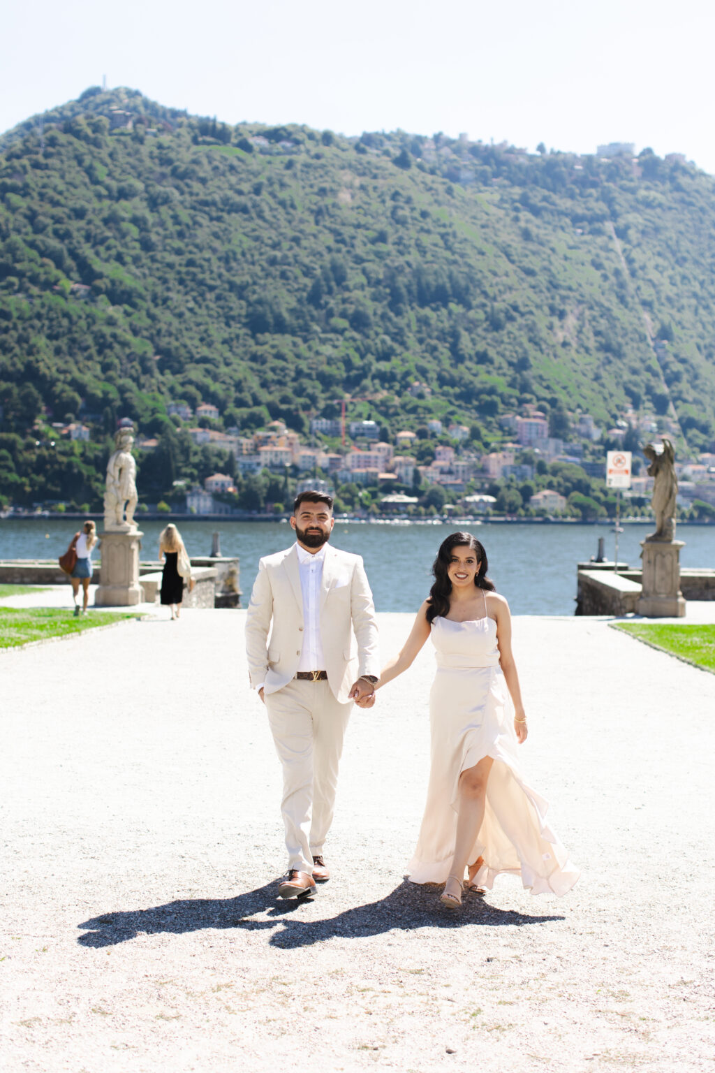 Couple walking hand in hand along a villa garden path flanked by statues toward the camera, Lake Como and the town of Como behind them