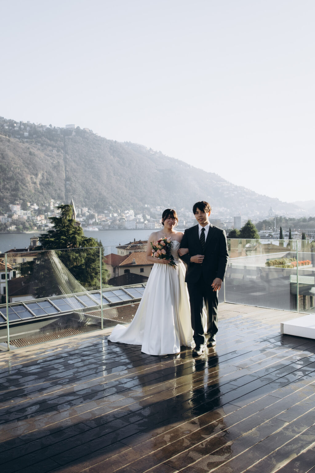Bride and groom walking arm in arm across a wet rooftop deck mirrored with reflections, Lake Como and the mountains behind them