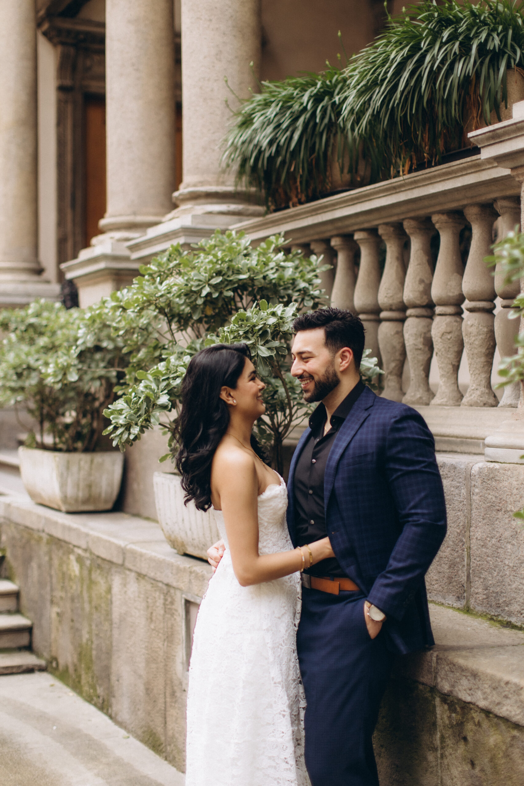 Courtyard Couple Photoshoot in Soft Daylight, Milan A couple in formal attire stands close together by a stone balustrade with greenery in a Milan courtyard in soft daylight