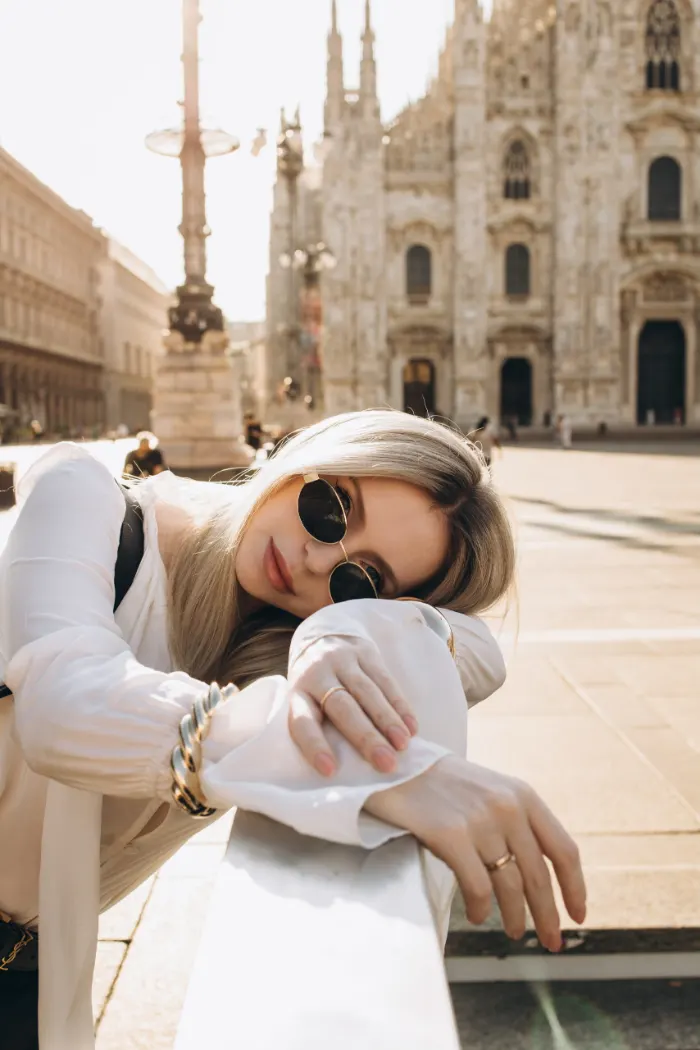 Close portrait of a blonde woman in round sunglasses resting her head on her arms along a white railing, with Milan Cathedral blurred behind her