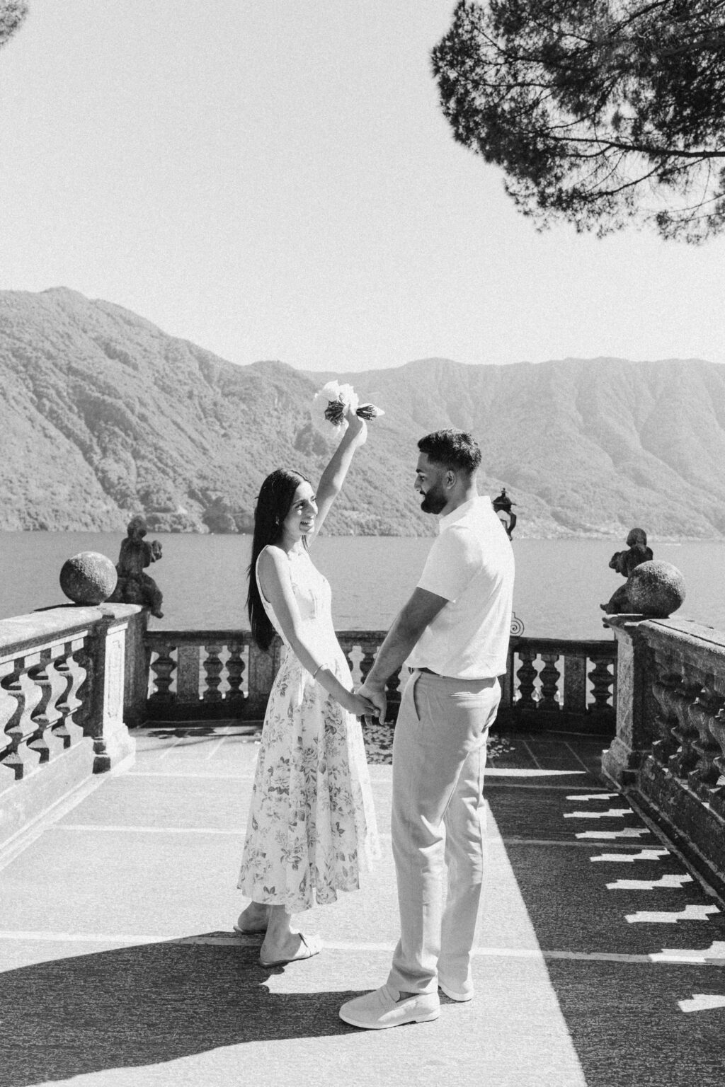 Black-and-white photo of the newly engaged woman raising her bouquet while holding her fiancé's hand on a terrace with stone cherubs above Lake Como